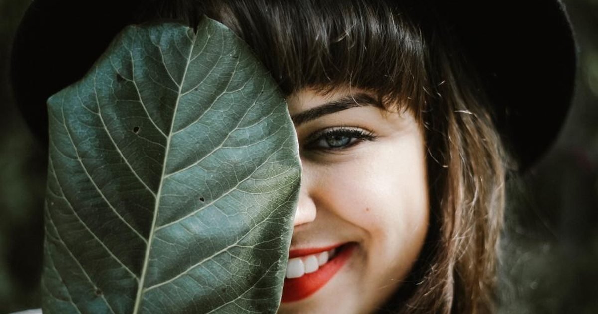 Smiling woman in a wide-brimmed hat holding a large leaf beside her face