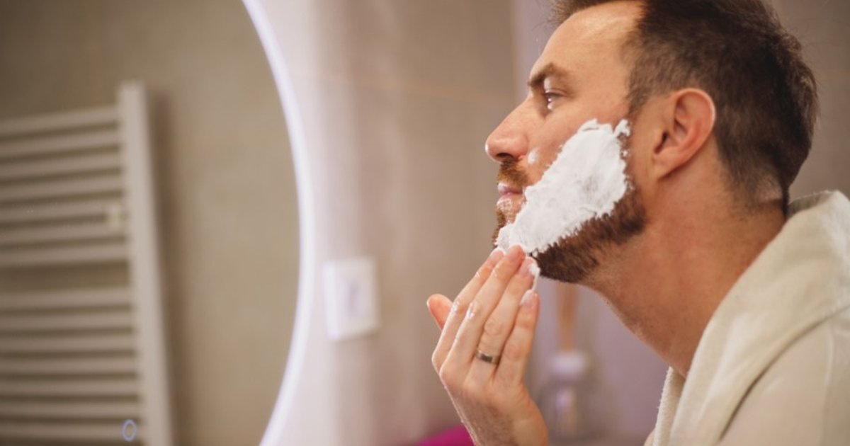 Man in a bathrobe applying shaving foam at a lit bathroom mirror