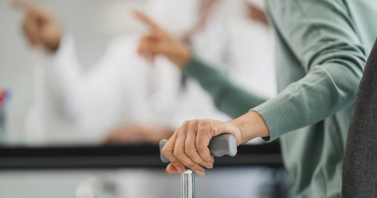 Older woman resting her hands on a walking cane while a clinician speaks nearby
