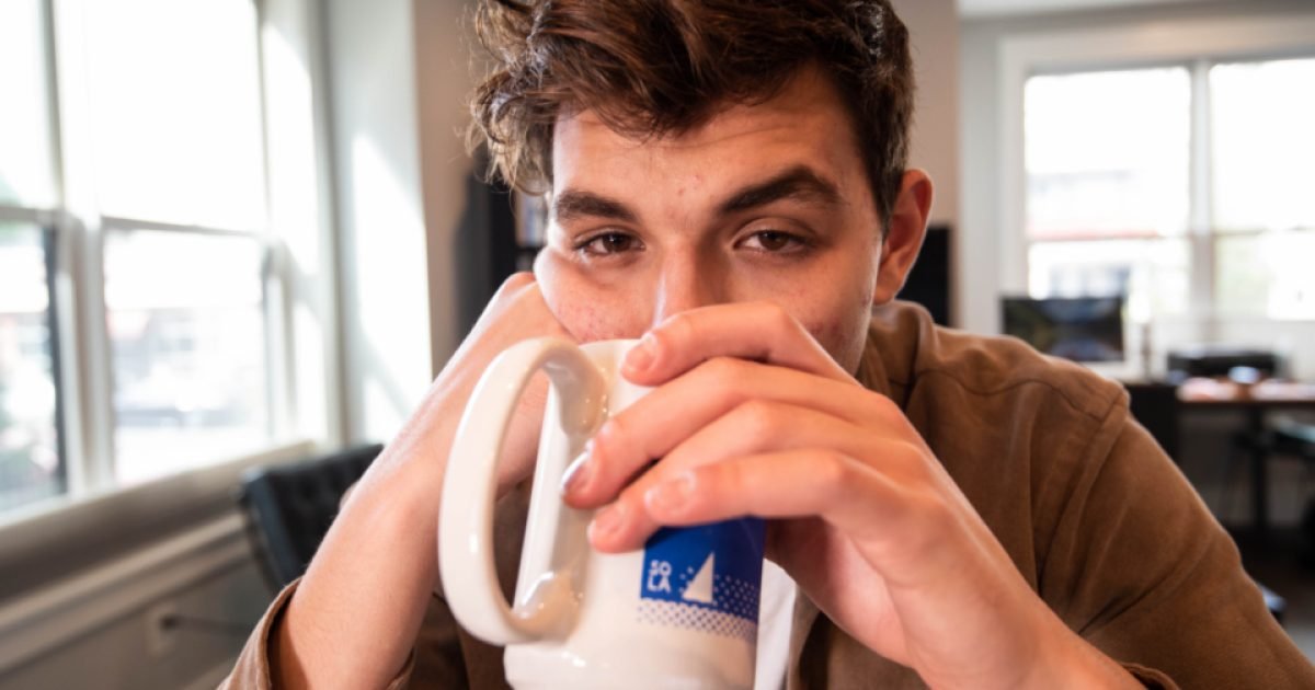 Young man drinking from a white mug at a table near a window