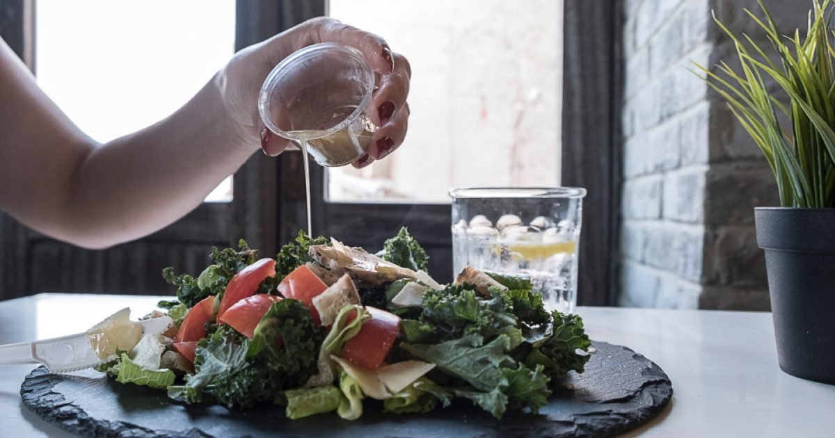 Person pouring dressing over a kale and tomato salad at a table