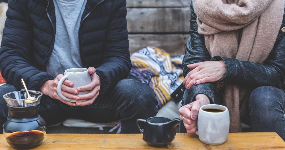Two people sitting outdoors holding mugs of coffee at a wooden table
