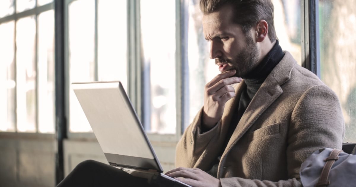 Man in a coat working on a laptop by a window with his hand on his chin