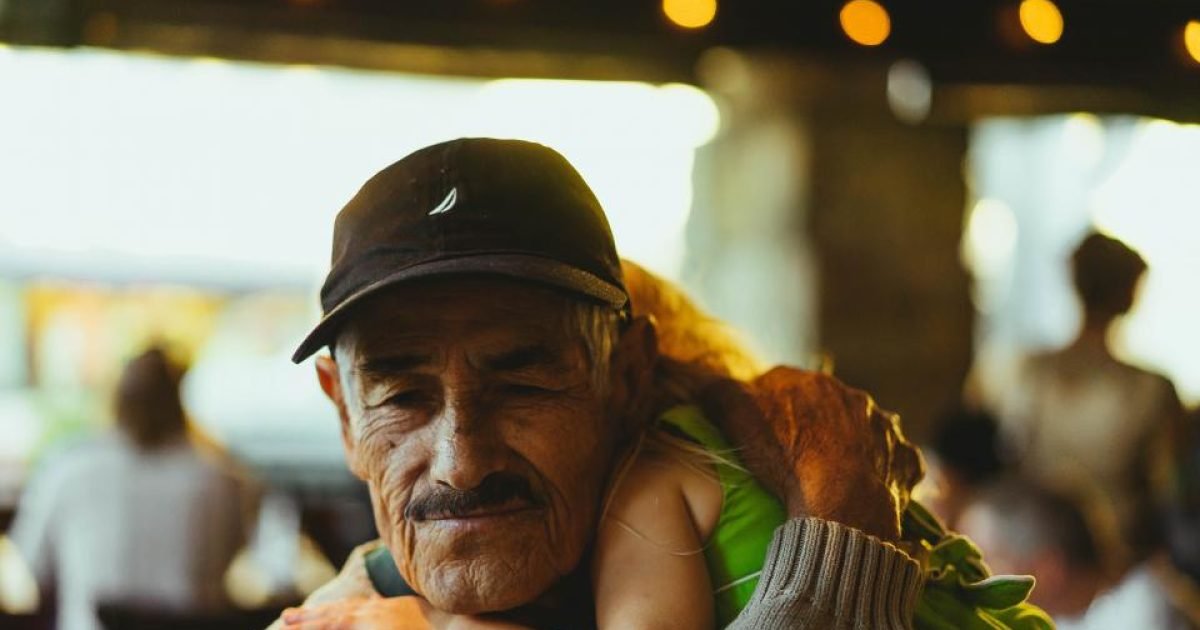 Older man in a cap sitting outdoors with a grandchild on his shoulders