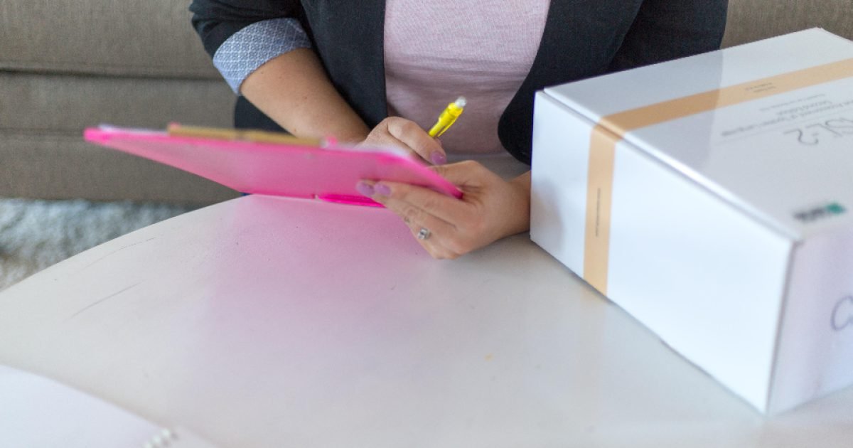 Woman sitting at a table writing a checklist on a clipboard at home