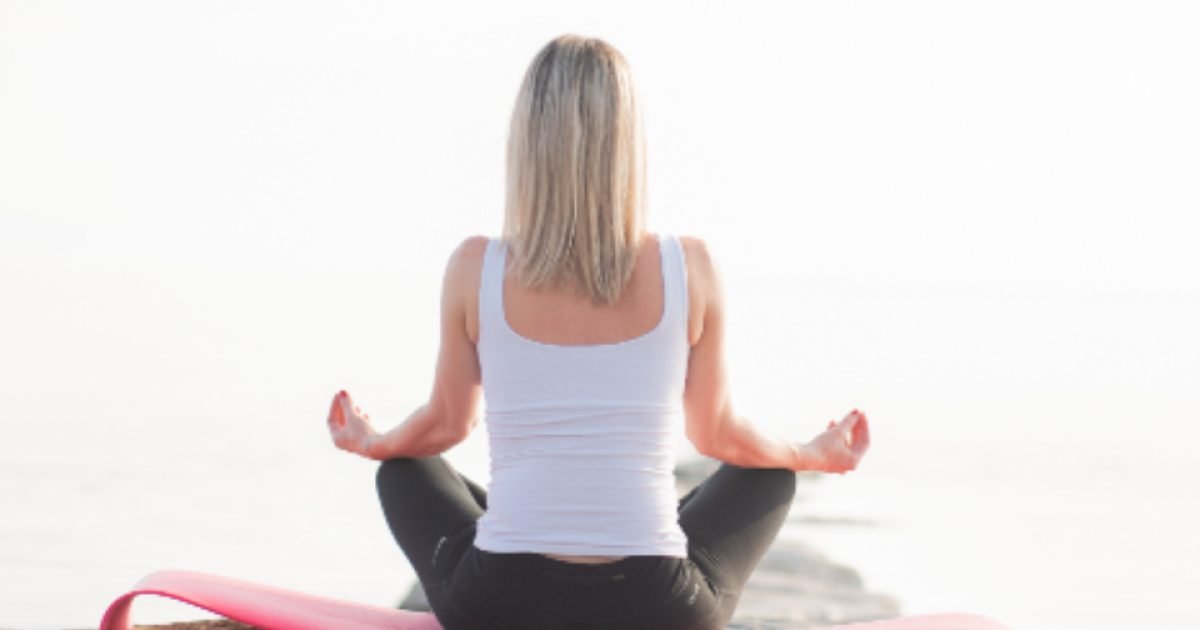 Woman sitting cross-legged on a yoga mat facing calm water at sunrise