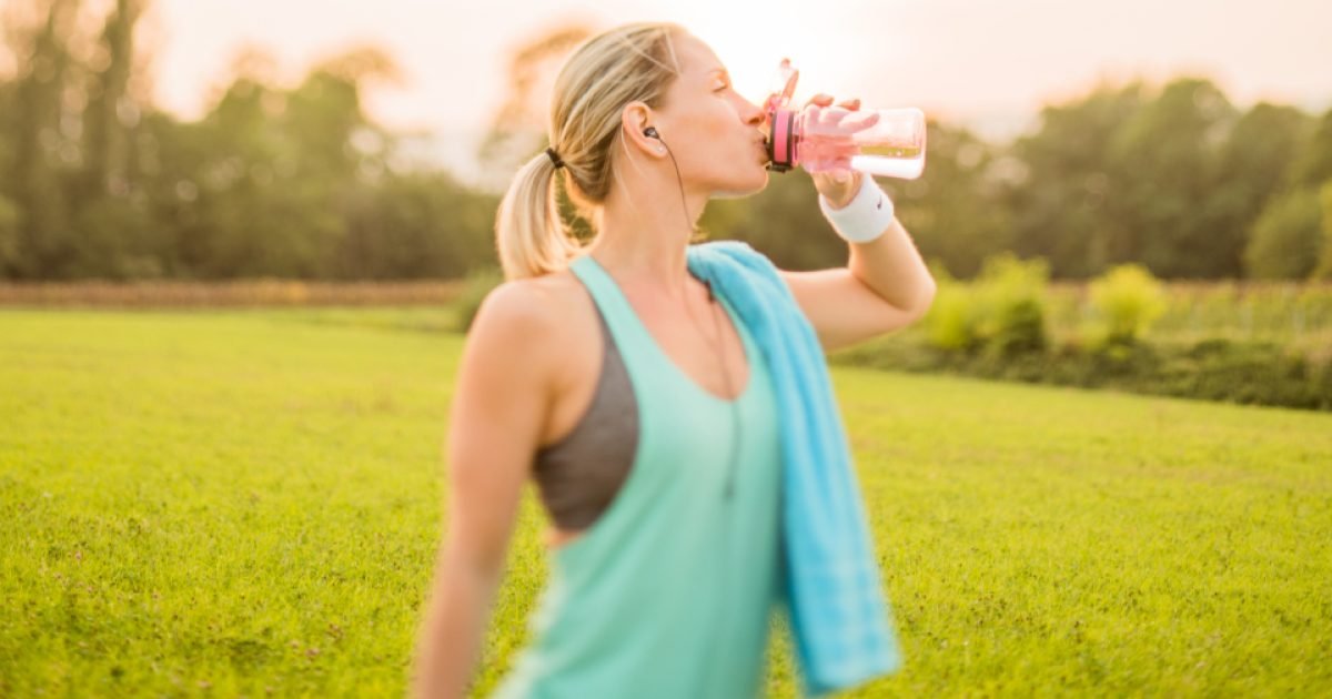 Woman in workout clothes drinking from a water bottle in a park at sunset