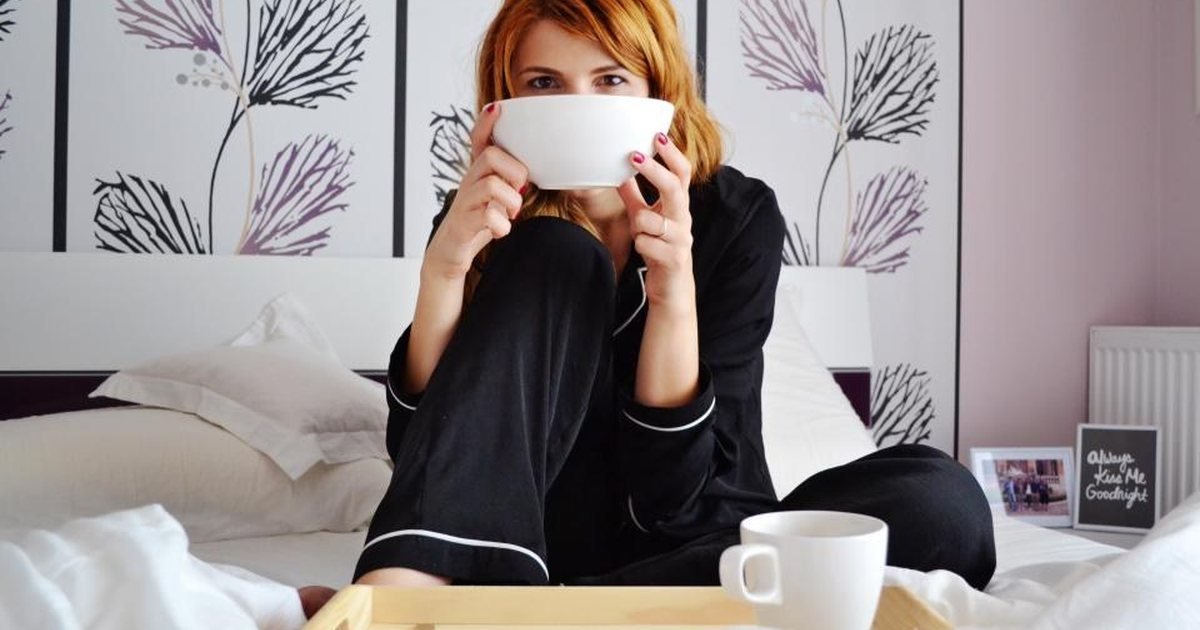 Woman sitting up in bed drinking from a cup beside a breakfast tray