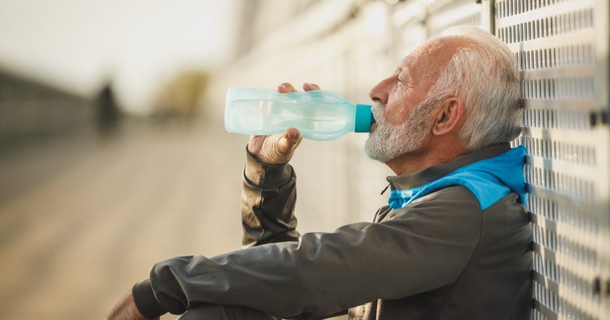Older man in a track jacket drinking water from a bottle while resting outdoors