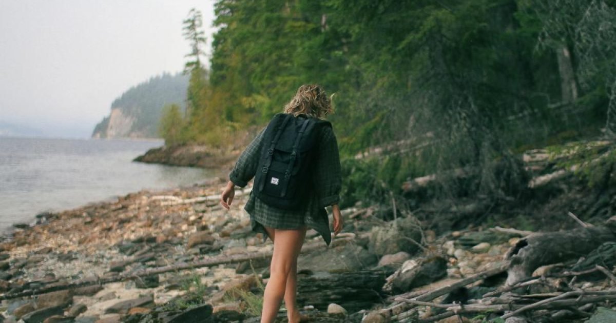 Woman with a backpack walking along a rocky shoreline beside a forest