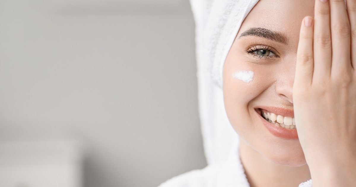 Smiling woman in a bathrobe and hair towel applying cream to her cheek